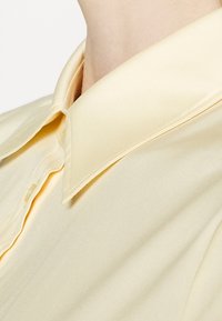 Gros plan d’une personne portant une chemise boutonnée jaune pâle avec un col pointu et une texture de tissu lisse.