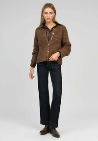 Gilet en tricot marron avec un col en V, agrémenté de grands boutons crème, associé à une chemise marron foncé et à un jean foncé évasé.