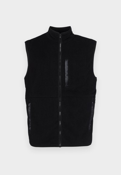 Melnā bezpiegu veste, kas izgatavota no mīksta flīsa, ar augstu apkakli, priekšējo rāvējslēdzēju un divām sānu kabatām, kā arī papildu krūšu kabatu.