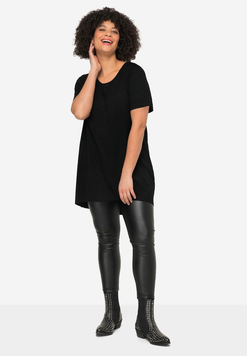 Femme souriante aux cheveux bouclés portant une tunique noire à manches courtes, des leggings en cuir noir et des bottines noires cloutées, debout sur fond blanc.