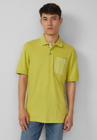 Jasnozielona koszulka polo z kołnierzykiem, krótkimi rękawami i kieszonką na piersi. Teksturowana tkanina, zapięcie na guziki i luźny krój.