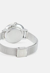 Reloj con correa de malla plateada y caja de acero inoxidable pulido, con un fondo texturizado. Presenta un cierre con un discreto grabado del logo.