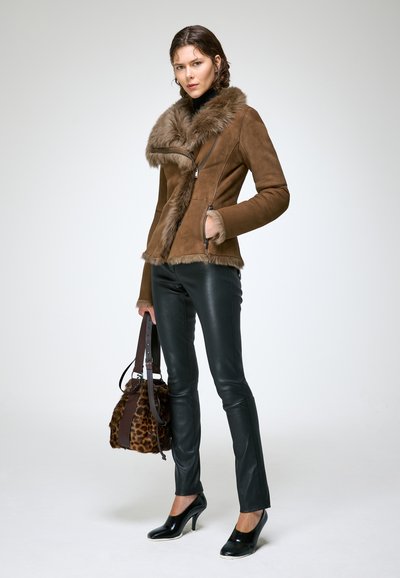 Femme portant une veste marron doublée en fourrure et un pantalon en cuir noir, tenant un sac à imprimé léopard, debout devant un fond blanc.