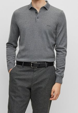 Mand iført en grå langærmet polo med "BOSS" logo og grå bukser, med sort bælte, hænder delvist i lommerne.
