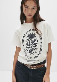 Ung kvinde iført en hvid t-shirt med sort blomster- og måne-design, parret med mørke jeans og et brunt bælte med metallæster.