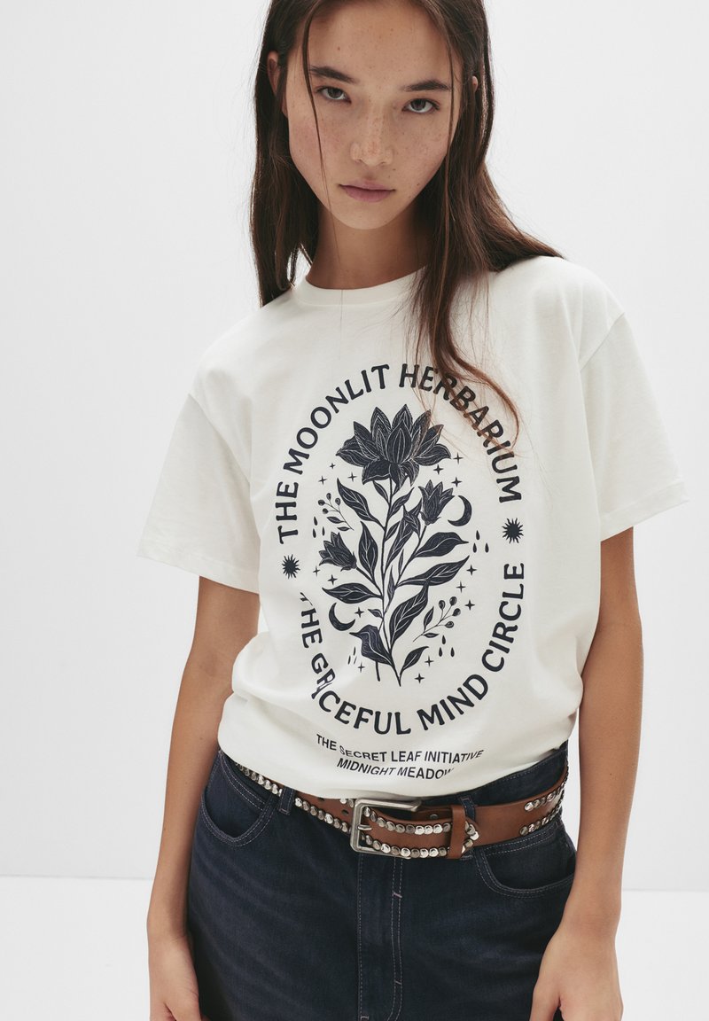 Ung kvinde iført en hvid t-shirt med sort blomster- og måne-design, parret med mørke jeans og et brunt bælte med metallæster.