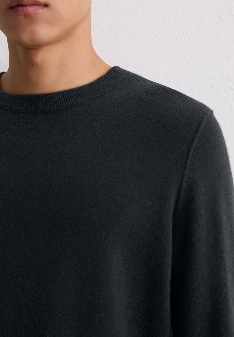 Ciemnoszary sweter wykonany z miękkiego materiału, z klasycznym okrągłym dekoltem i ściągaczem. Tekstura wygląda na gładką i drobno dziewiarską.