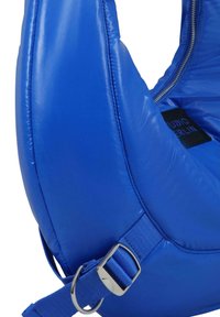 Blå vadderad stol med en elegant böjd design, utrustad med en dragkedja, textiletikett och justerbar rem med ett metallspänne.