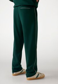 Groene trainingsbroek met een rechte snit, voorzien van witte piping langs de naden en een achterzak. Gecombineerd met beige en groene sneakers.