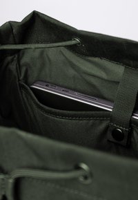 Borsa tote in tessuto verde con chiusura a coulisse. Tasca interna per smartphone, che mostra texture lisce e opache e un hardware minimale.