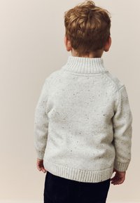 Lysegrå strikket sweater med høj ribbet krave og plettet tekstur, kombineret med mørke jeans. Set fra bagsiden.