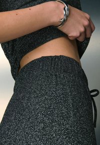 Sort metallisk strikbluse og matchende bukser med elastisk taljebånd, fremhævet med et skinnende sølvarmbånd på en bar arm. Glat tekstur og tætsiddende design.