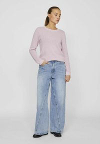 Ljuslila texturerad stickad tröja med långärmar, kombinerad med vida ljusblå jeans. Modellen står med ena handen i fickan.