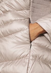 Vadderad, ljusbrun ytterklädsel med räfflad krage och dragkedjeficka. Slät, glänsande tyg med diagonala sömdetaljer.