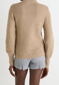 Beżowy sweter wełniany z wysokim kołnierzem i prążkowanym wzorem, w połączeniu z jasnoszarymi, dzianinowymi szortami z wzorem w warkocze na końcach.