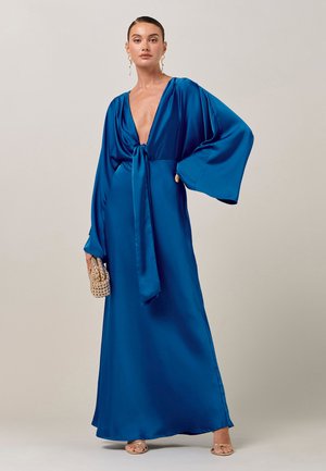 Nainen, joka pukeutunut syvän siniseen satiinimekkoon, jossa on pitkät piispan hihat, V-pääntie ja solmittava etuosa, kultaiset sandaalit, kultaiset korvakorut ja kultainen helmikoristeltu iltalaukku kädessään.