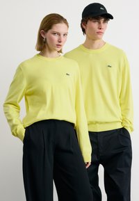 Δύο μοντέλα φοράνε ταιριαστά κίτρινα πουλόβερ Lacoste και μαύρο παντελόνι, το ένα με κοντά μαλλιά, το άλλο με μαύρο καπέλο, στέκονται μπροστά σε λευκό φόντο.