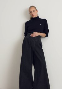Maglione nero a costine con collo alto abbinato a pantaloni in denim neri a gamba larga e vita alta. Design minimalista con un sottile dettaglio del logo.