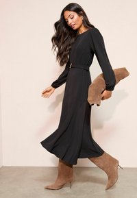 Dlouhé černé šaty s dlouhými rukávy a zúženým pasem, doplněné béžovými semišovými kotníkovými botami. Látka se volně spouští až k podlaze.