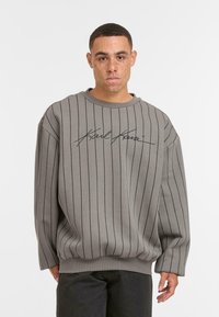 Giovane uomo con capelli corti e lentiggini, che indossa una felpa grigia a righe con testo corsivo nero e pantaloni scuri, in piedi su uno sfondo bianco.