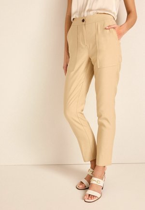 Frau trägt beige Hose mit hoher Taille und großen Vordertaschen, kombiniert mit weißen Sandalen mit offenen Zehen und goldenen Schnallen.