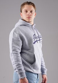 Grauer Kapuzenpullover mit Fronttasche und gesticktem Text. Verfügt über gerippte Manschetten und Saum. Kombiniert mit Jeans.
