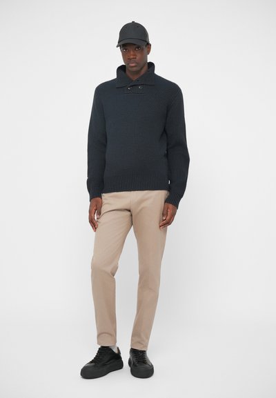 Tummansininen neulepusero korkean kauluksen ja kahden napin kanssa, yhdistettynä beigeihin housuihin ja mustiin lenkkitossuihin. Lippis samaa väriä.