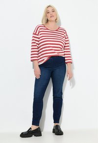 Žena stojící proti bílé zdi, oblečená v červeno-bílém pruhovaném tričku s dlouhým rukávem, tmavě modrých skinny džínách a černých mokasínách.