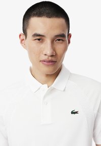 Vit pikétröja med krage och tre knappar, med texturerade detaljer på ärmarna och en grön logotyp på bröstet.