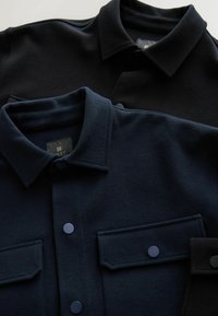 Zwei Hemden mit Knopfleiste, eines in Schwarz und eines in Dunkelblau, verfügen über einen Kragen, Brusttaschen und sichtbare Nähte auf einem strukturierten Stoff.