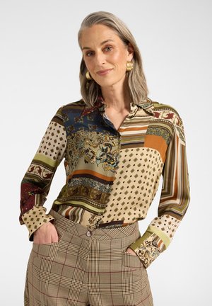 Donna con capelli grigi indossa una camicetta colorata a fantasia e pantaloni a vita alta a quadri con le mani nelle tasche, sorridendo dolcemente su sfondo bianco.