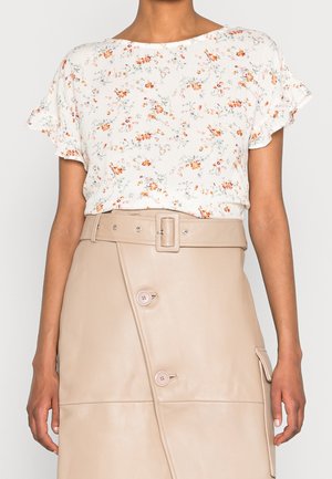 Kobieta w białej, kwiecistej bluzce z krótkim rękawem, włożonej w beżową skórzaną spódnicę z guzikami, paskiem i boczną kieszenią.