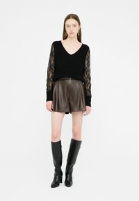 Maglione nero a maglia con scollo a V e maniche lunghe in pizzo, abbinato a una gonna di ecopelle marrone e stivali neri con tacco al ginocchio.