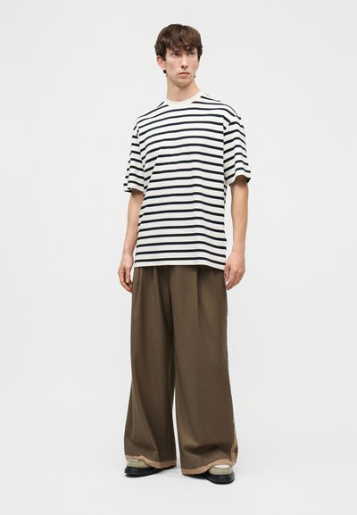 Νέος άνδρας που στέκεται, φοράει μπλούζα με κοντά μανίκια σε ασπρόμαυρες ρίγες, φαρδιά καφέ παντελόνια και μπεζ παπούτσια, μπροστά σε μια λευκή απλή επιφάνεια.