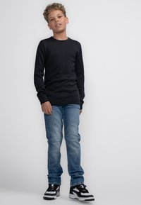 Zwarte trui, lichtblauwe spijkerbroek en zwart-witte sneakers. De trui heeft een ronde hals en lange mouwen, met ribbels aan de manchetten en onderkant.