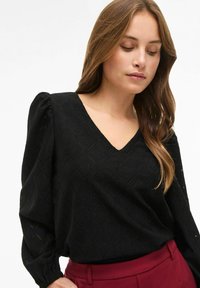 Blusa nera in tessuto strutturato con motivo a diamante, scollo a V e maniche a sbuffo. Abbinata a pantaloni bordeaux.