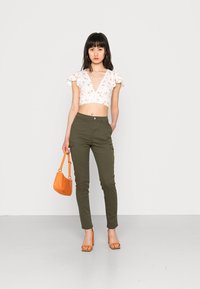 Biely kvetinový crop top s volánovými rukávmi, olivovo zelené cargo nohavice, oranžová kabelka a oranžové sandále na podpätku. Jemné textúry po celom oblečení.