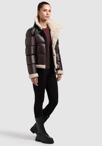 Giubbotto puffo marrone con colletto e polsini in shearling, dotato di zip frontale e dettagli in pelle nera; abbinato a leggings neri e stivali da combattimento.