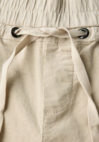 Pantalons en velours côtelé beiges avec une surface texturée, dotés d'une taille élastique et d'un cordon de serrage avec des accents métalliques noirs. Comprend une poche latérale.