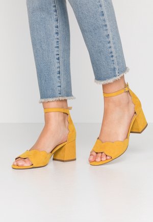 Yellow suede block-heeled sandals with scalloped edges, open toe design, and ankle strap. Paired with light-wash frayed hem jeans.