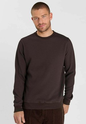 Man met kort haar en baard, die een donkerbruine gestructureerde trui met lange mouwen en bijpassende broek draagt, staand tegen een effen achtergrond.