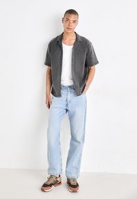 Camisa de botones de manga corta gris sobre una camiseta sin mangas blanca, combinada con jeans azul claro y zapatillas negras, naranjas y grises.