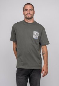 T-shirt en coton vert foncé avec un col rond et des manches courtes. Présente un design de poche imprimé en blanc et bleu.