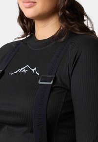 Black ribbed long-sleeve shirt with a white mountain graphic and adjustable black suspenders featuring embossed branding. Close-up view.