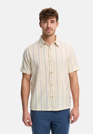 Mann trägt beige kurzärmliges Hemd mit dünnen blauen vertikalen Streifen und blaue Hose, steht vor einem einfarbigen weißen Hintergrund.