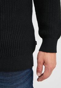 Schwarzer Strickpulli mit einem strukturierten Muster, gerippten Bündchen und geradem Saum. Sichtbare Hand, die die Seite des Pullis hält.