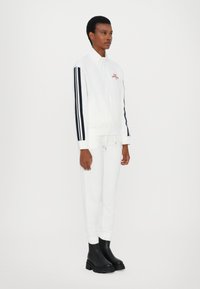 Witte sportieve trainingspak met marineblauwe strepen op de mouwen, bestaande uit een ritsjack en joggingsbroek met koordjes, gecombineerd met zwarte enkellaarsjes.