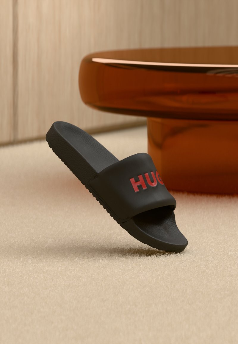 Sandale noire à semelle plate avec logo "HUGO" rouge sur la bride, positionnée en angle sur un tapis beige à côté d'un pied de table rond marron brillant.