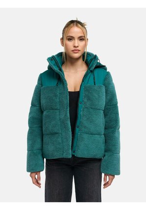 Kvinde iført teal dunjakke med fleece tekstur, hætte, sort top og mørke jeans, stående mod en hvid baggrund.