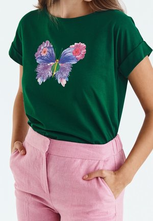 Nainen pukeutuneena tummanvihreään t-paitaan, jossa on perhonen, joka on tehty vaaleanpunaisista ja purppuraisista kukista, yhdistettynä korkeavyötäröisiin vaaleanpunaisiin housuihin, kädet taskuissa.